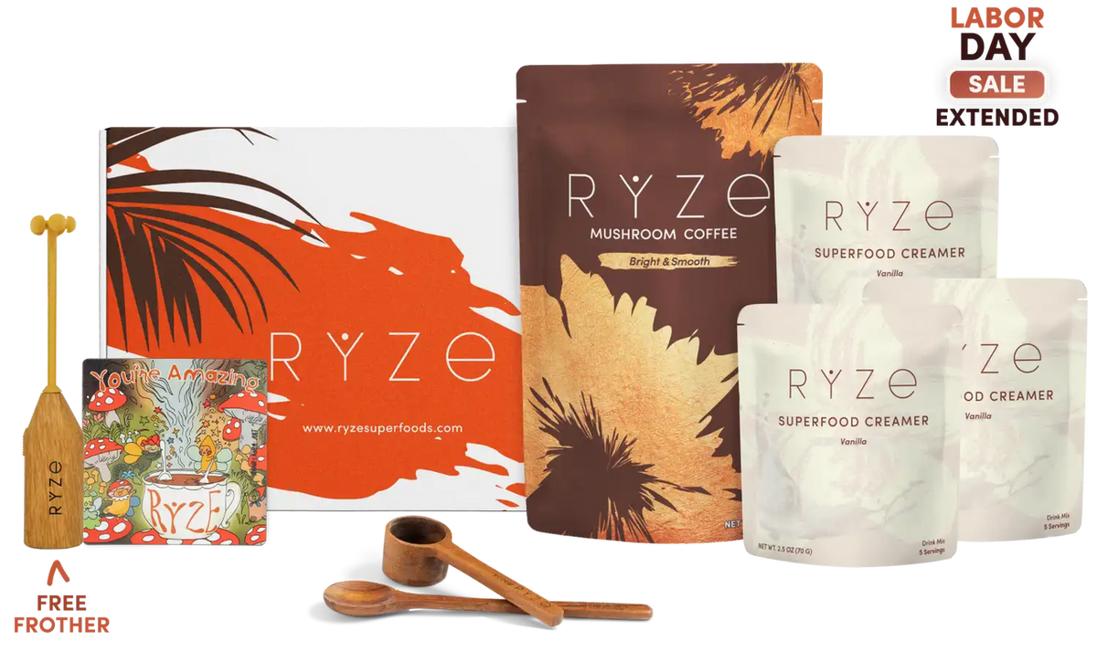Ryze mushroom coffee gift set featuring a large orange and white box, a brown pouch labeled 'RYZE MUSHROOM COFFEE', three white pouches of 'RYZE SUPERFOOD CREAMER' in vanilla, coconut, and chocolate flavors, two wooden measuring spoons, a wooden spoon handle, and a 'FREE FROTHER' gift prop, with a 'LABOR DAY SALE EXTENDED' badge.