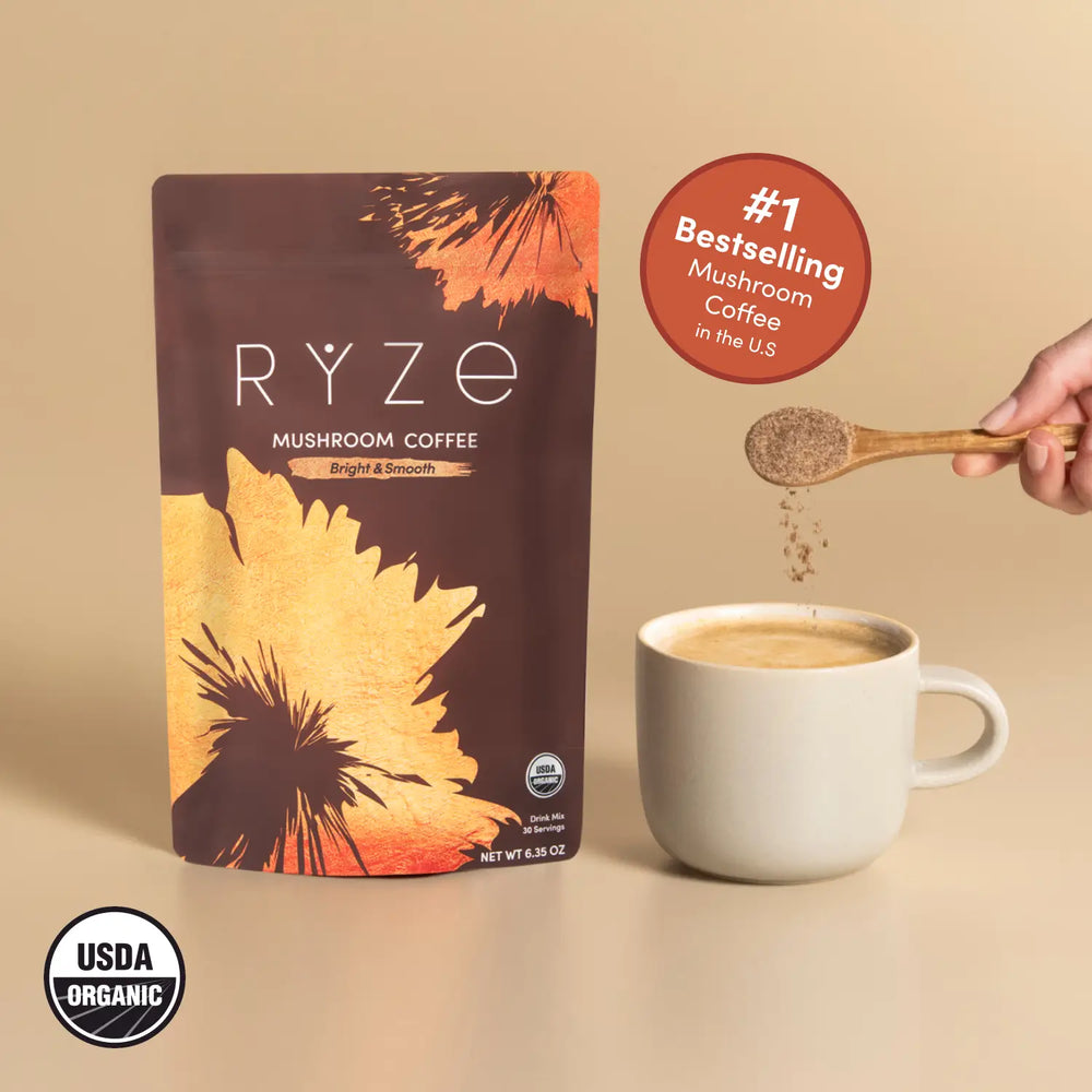 A stand-up pouch of RYZE Mushroom Coffee stands on a beige surface beside a white mug with coffee and a hand holding a wooden spoon sprinkling powder. A red badge says '#1 Bestselling Mushroom Coffee in the US' and a USDA Organic seal appears in the lower left.